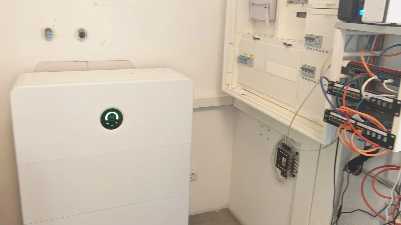 Batteriespeicher mit leuchtender grüner Statusanzeige neben einem geöffneten Zählerschrank mit Verkabelung