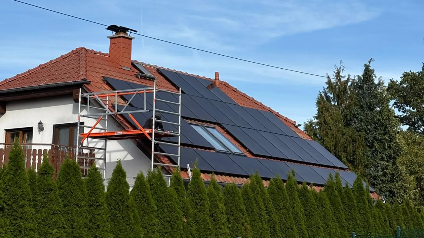 Handwerker montiert Solarmodule auf einem Satteldach, im Vordergrund ein Gerüst, eine Dachhälfte ist bereits vollständig mit schwarzen Modulen belegt