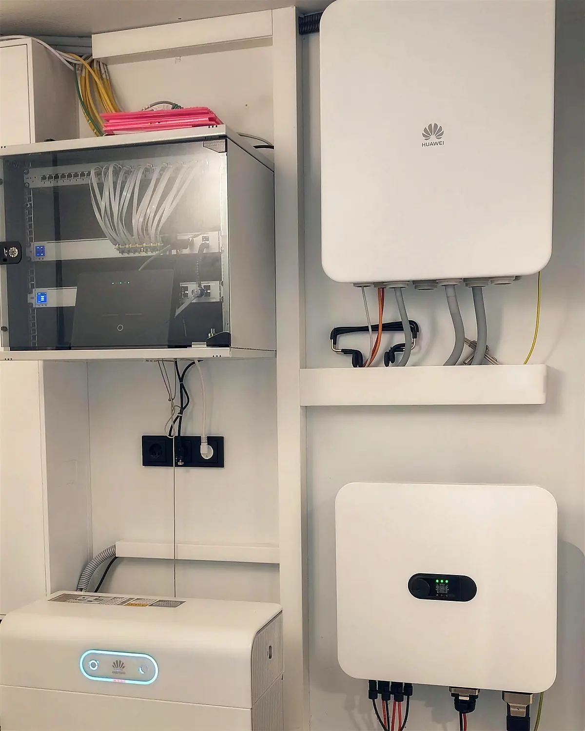 Technikraum mit zwei wandmontierten Wechselrichtern, Batteriespeicher, Netzwerkschrank mit sortierten Patchkabeln und sauber geführten Leitungen